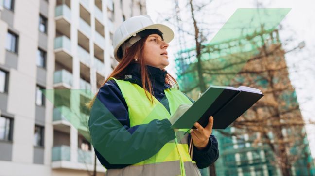 A female access consultant in a hardhat and high visibility vest is standing outside a large building and taking notes with VAC Green tick