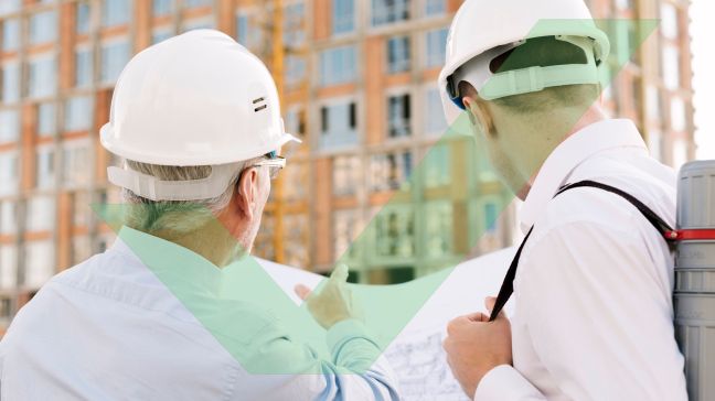 Two male access consultants are standing outside and wearing hardhats while looking at an architectural drawing, one is pointing at the construction site with VAC Green tick